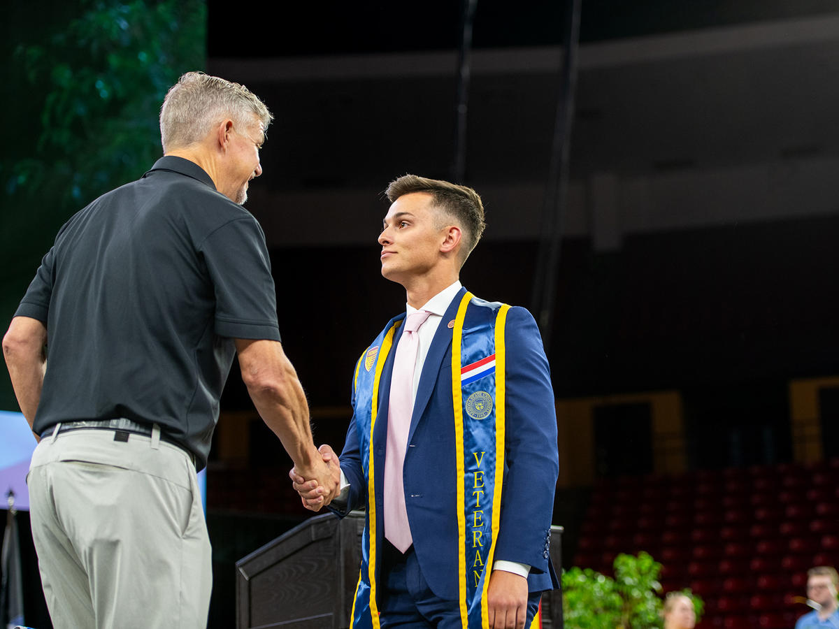 Two people shaking hands at a graduation event, one wearing a blue suit and veteran sash.